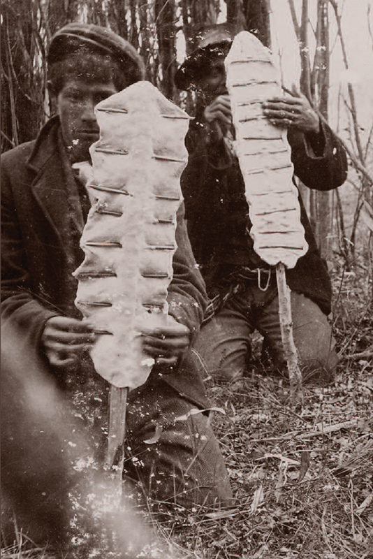 Historic black-and-white photo of two men each showing a long, thin piece of dough stretched out with sticks.