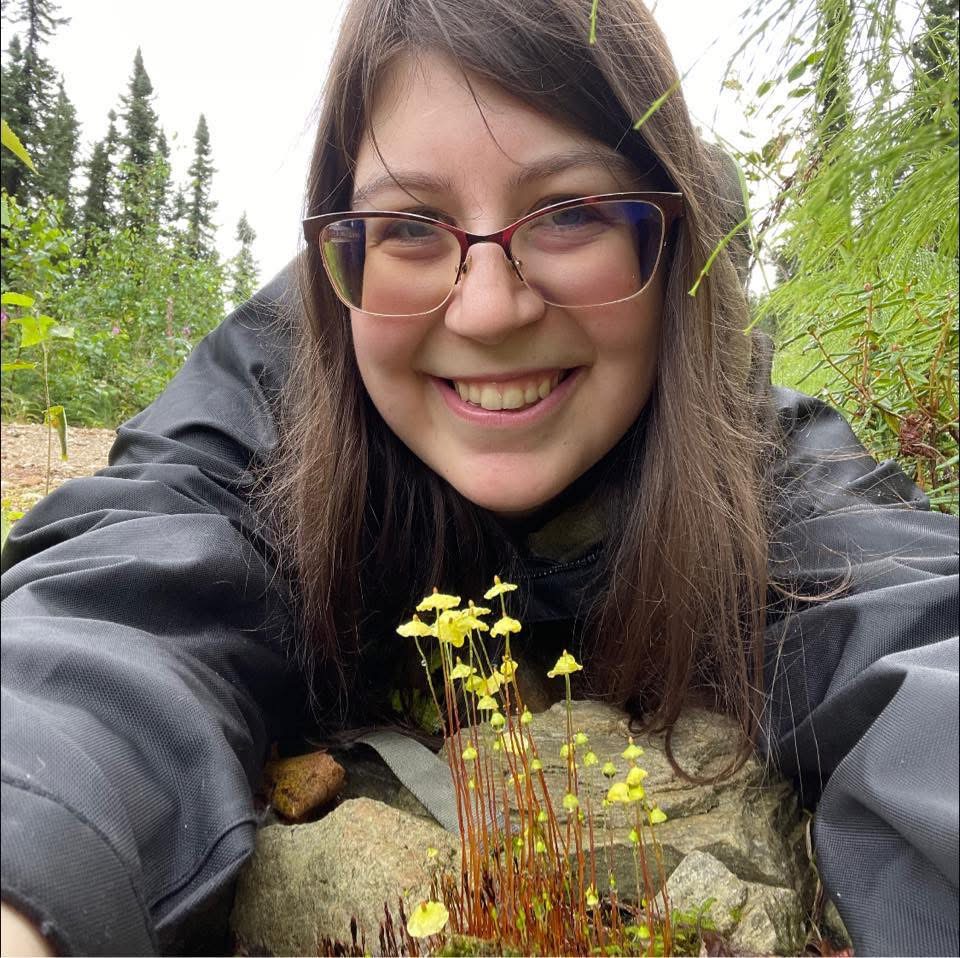 Portrait of young woman with long, brown hair. Tuft of moss in foreground